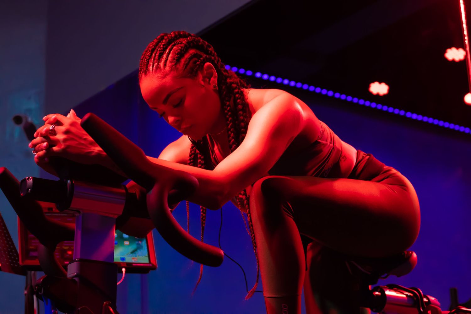 Performer in red lighting crouches dramatically on stage on a cycling bike during an energetic work out session.