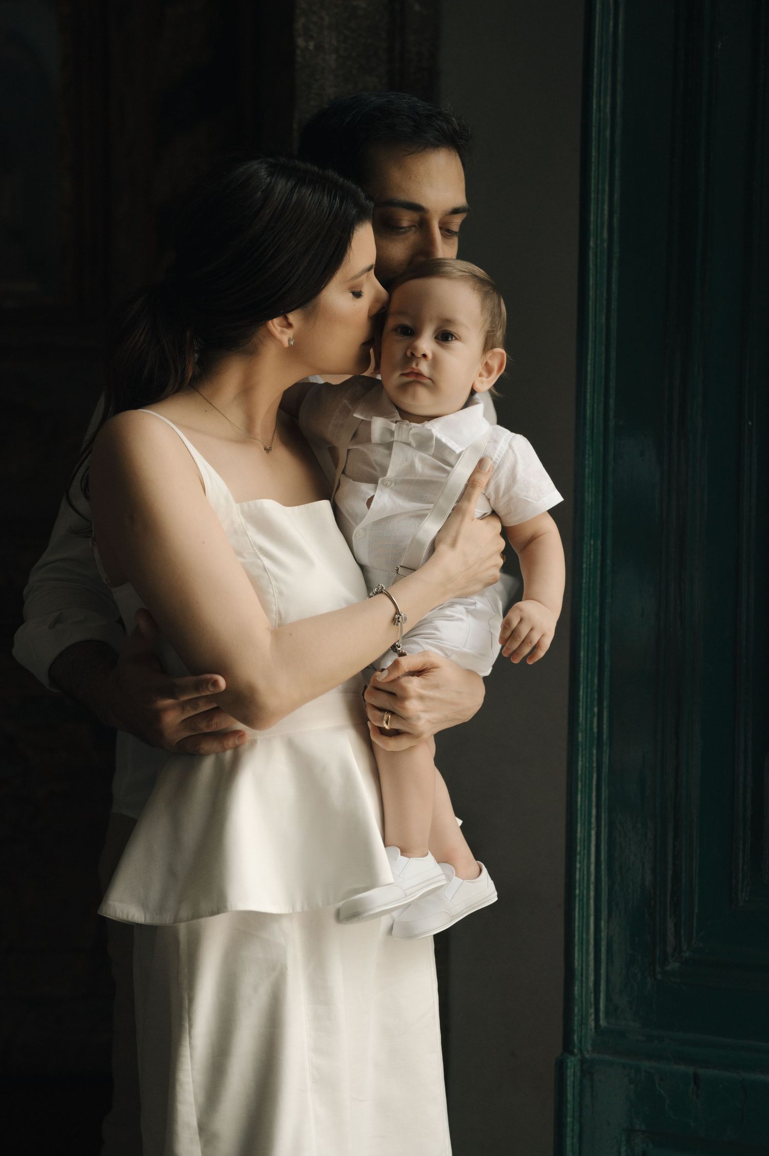 A tender moment between parent and baby wearing white outfits against a dark background in a series of intimate portraits.