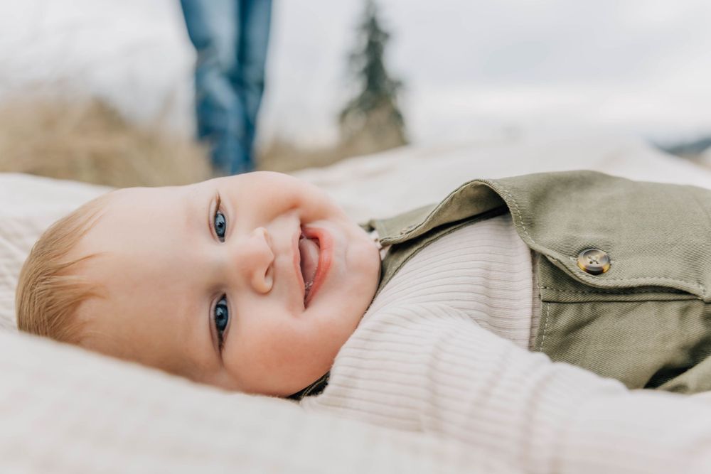 Smiling baby with blue eyes lying on a white blanket outdoors wearing a green outfit.