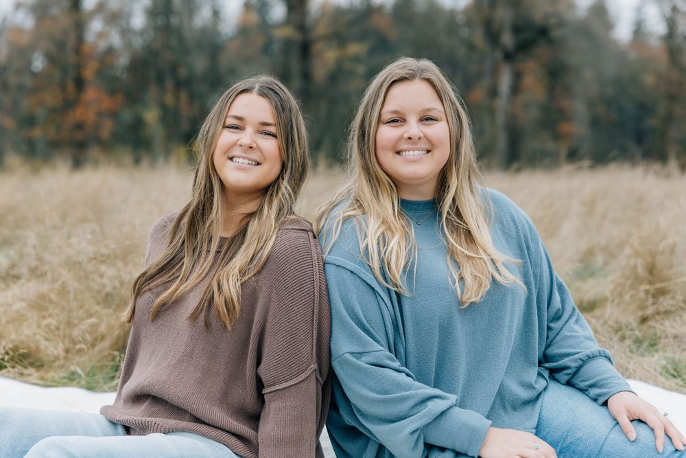 Two young blonde women smiling together seated in a golden autumn meadow.