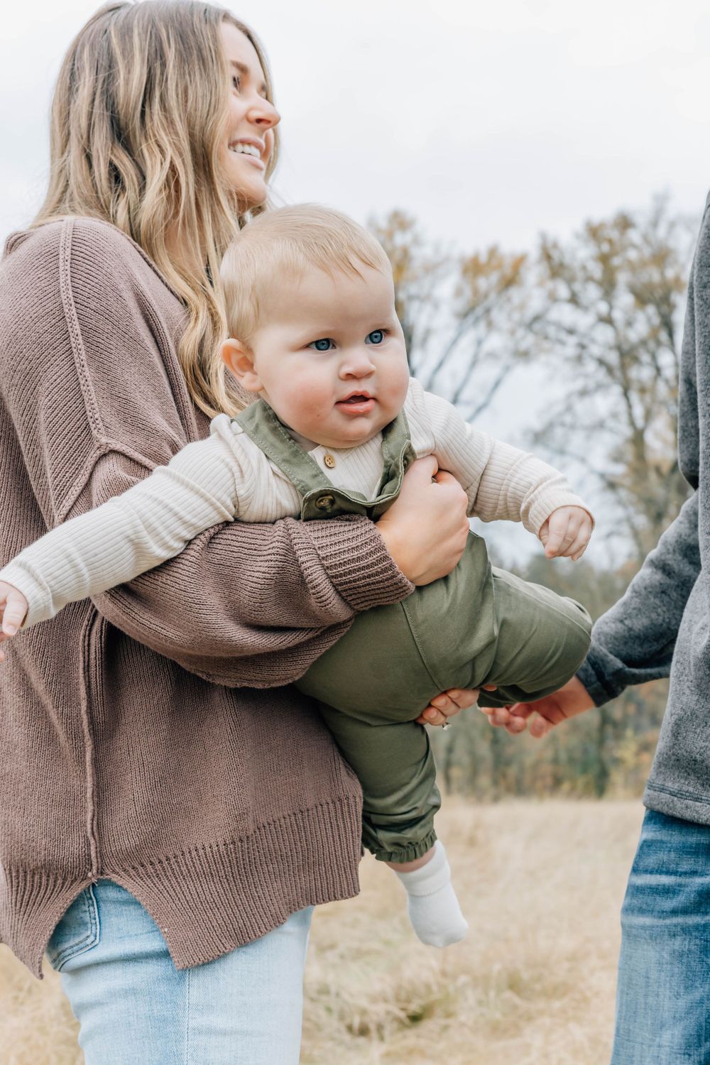 Mother holding a smiling baby in a green outfit outdoors in an autumn field.