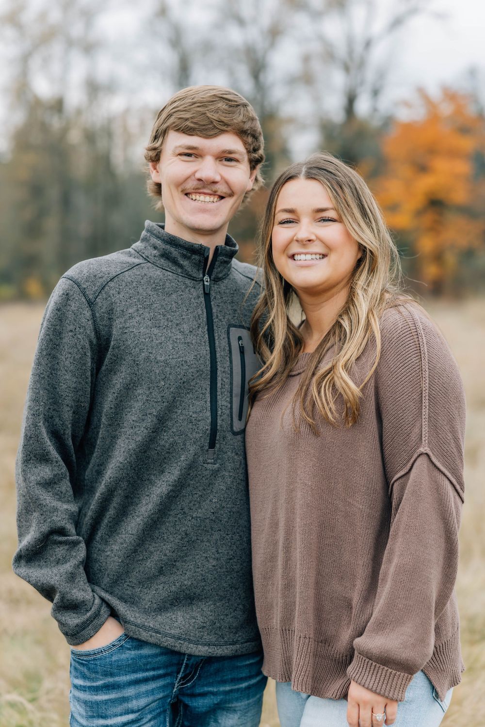 Smiling young couple posing together in a fall outdoor setting with autumn foliage.