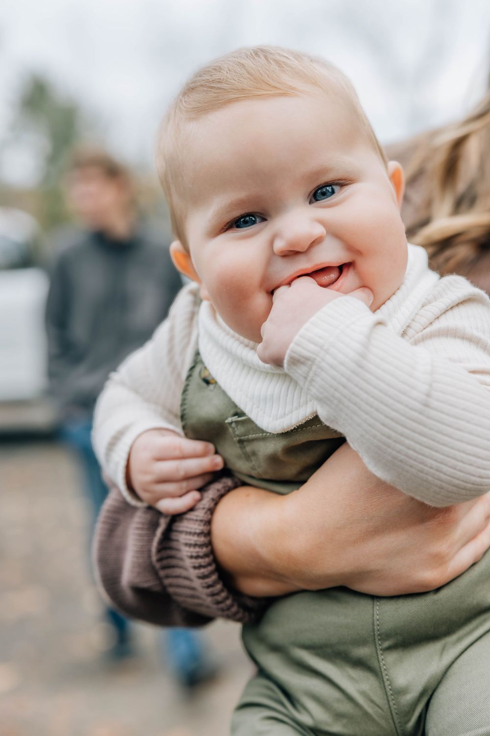 Adorable blue-eyed baby smiling with hand in mouth being held outdoors.