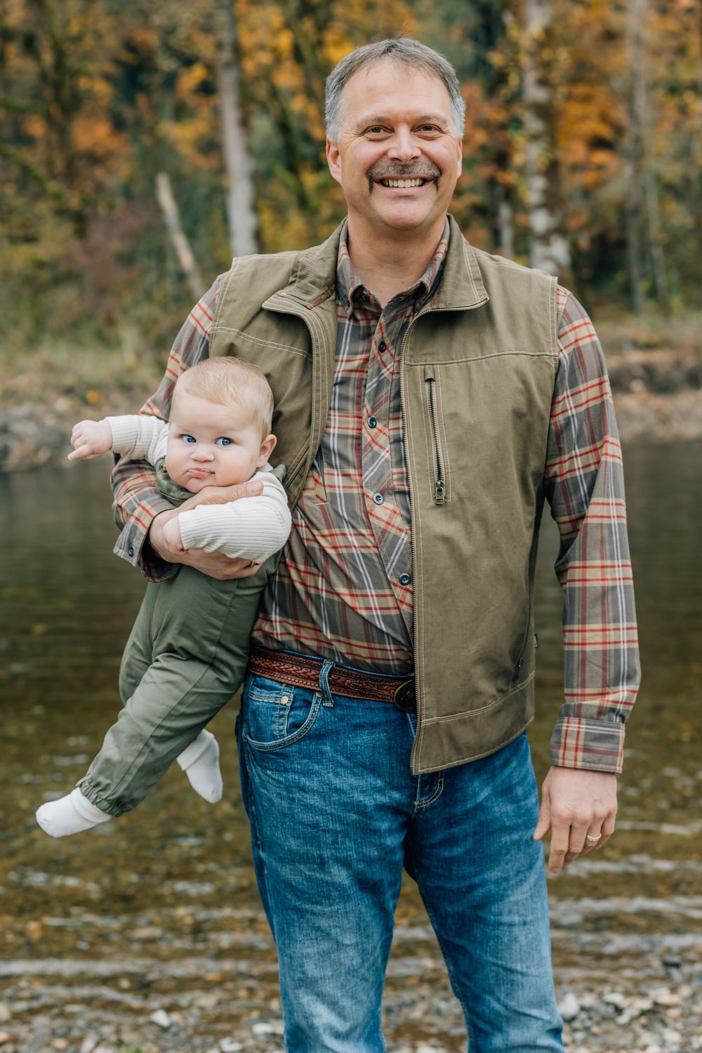 Smiling man holding a baby outdoors near a river with colorful autumn foliage in the background.