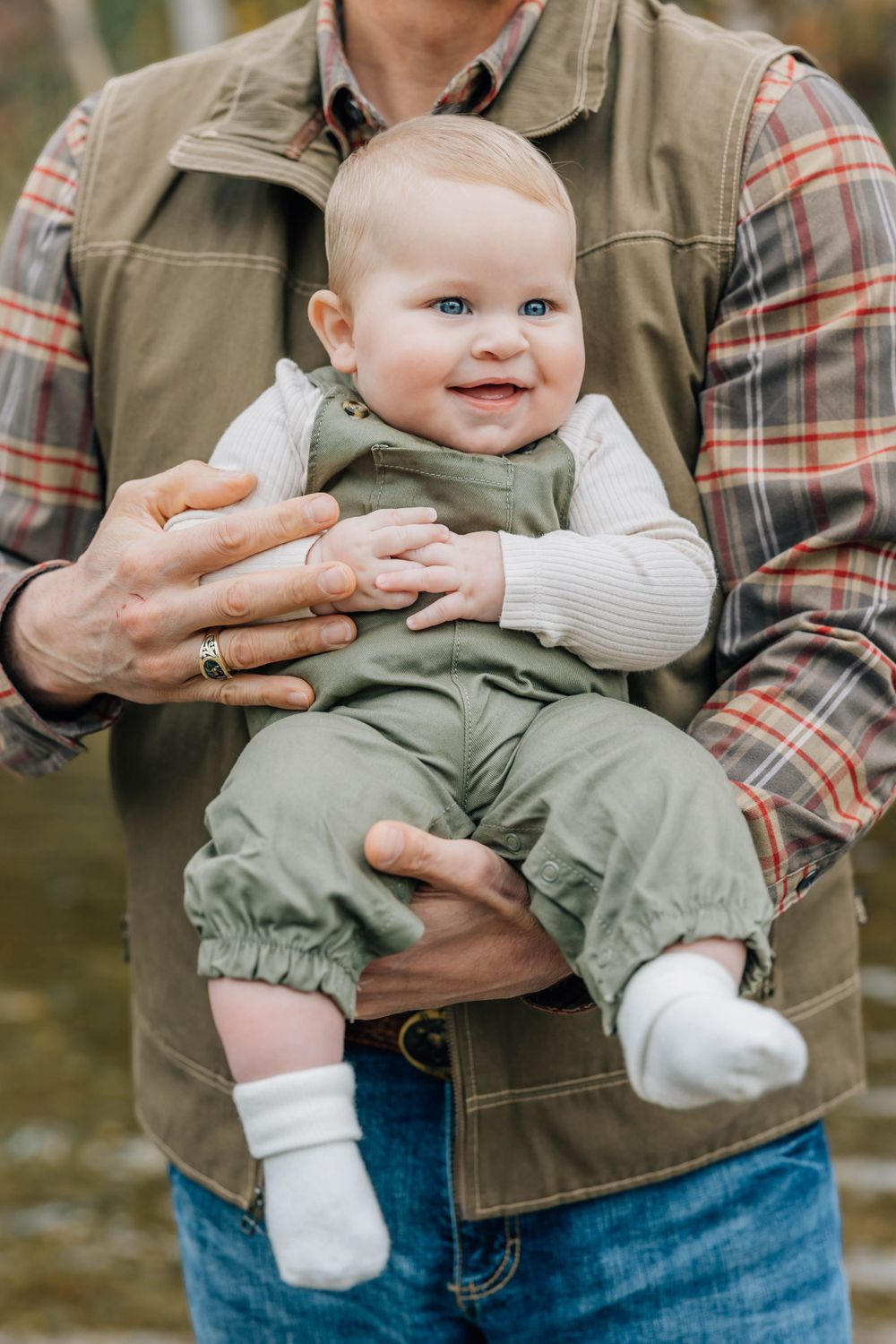 Cheerful blue-eyed baby in green outfit being held by an adult outdoors.