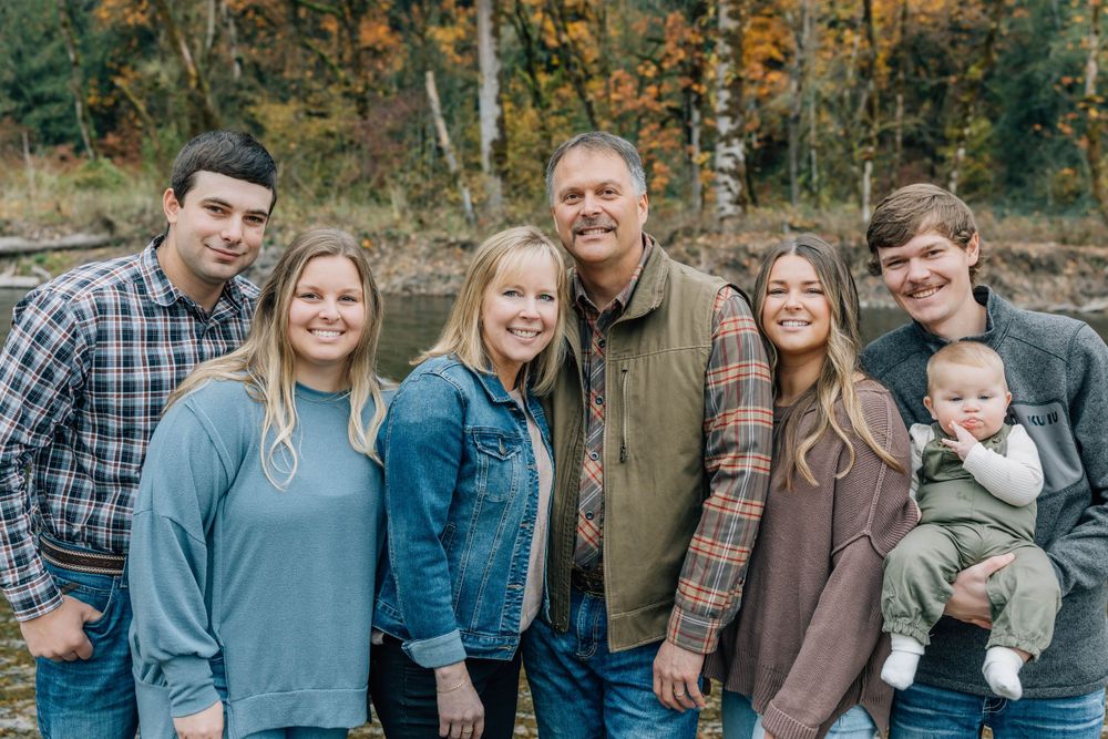Family of seven smiling together outdoors with vibrant fall foliage in the background.