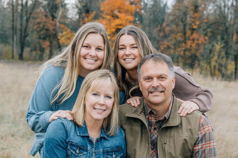 Family of four smiling together in an autumn field with colorful fall trees behind them.