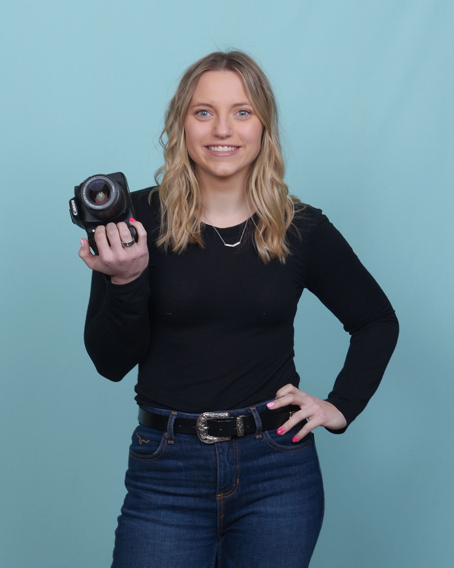 Photographer wearing a black sweater and jeans holds a professional camera against a light blue studio background.
