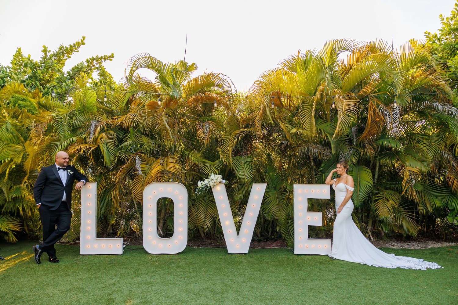 Wedding couple and guests pose with large white LOVE letters in front of tropical palm trees during outdoor celebration.