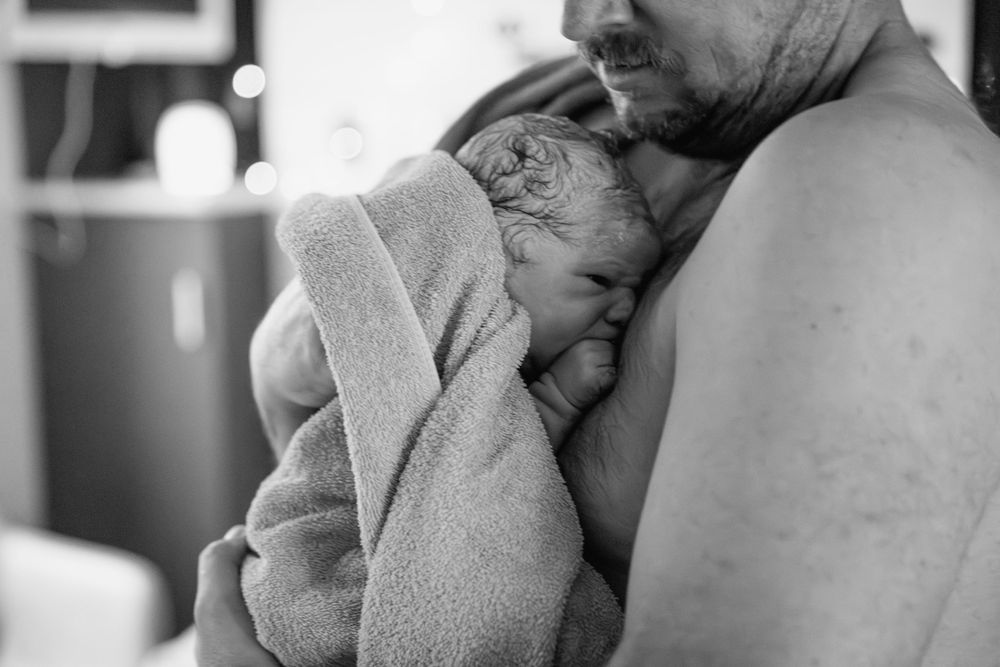 A tender black and white moment showing a newborn swaddled in a soft towel being held close against his dads chest.