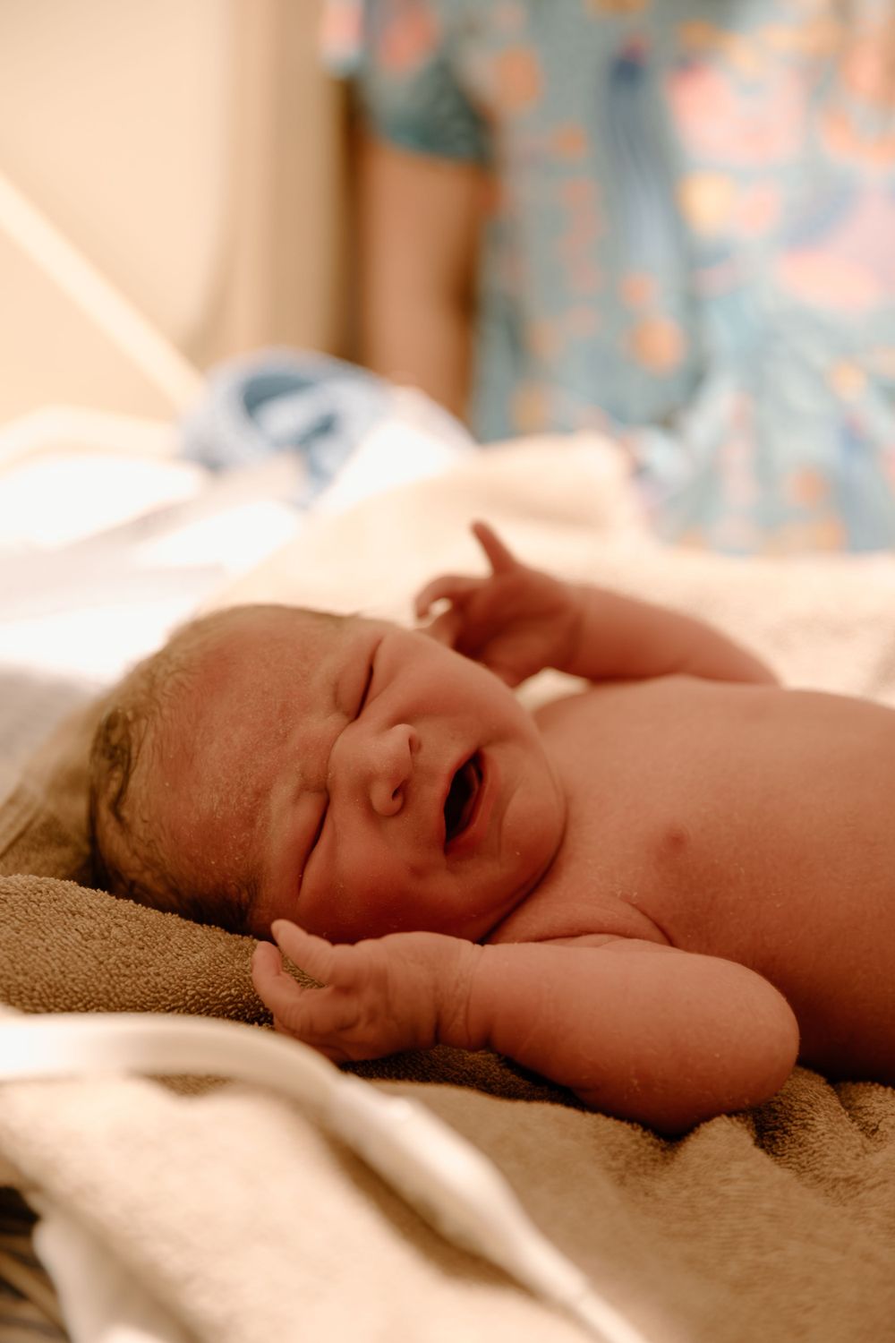 A crying newborn lies on a cream blanket in warm lighting.