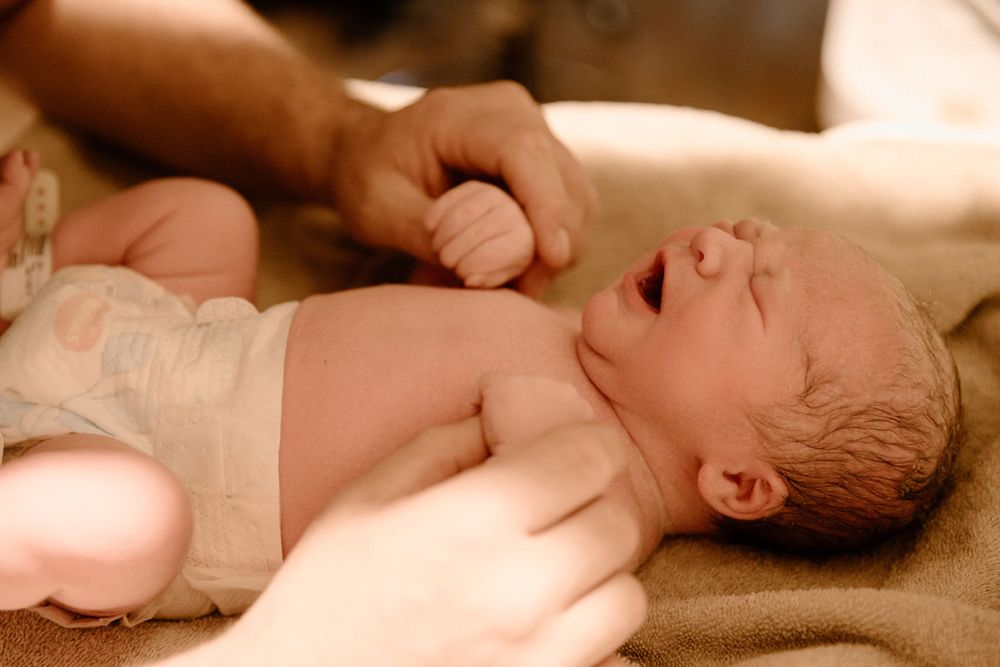 A crying newborn baby lies on a soft brown blanket while being comforted by gentle hands.
