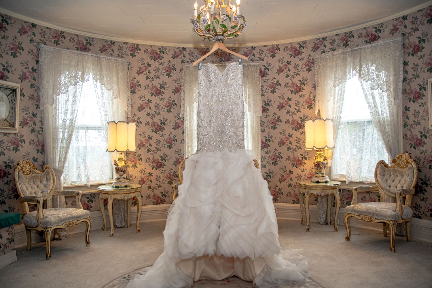 Wedding dress hangs in floral wallpapered room with antique furniture and lace curtains.