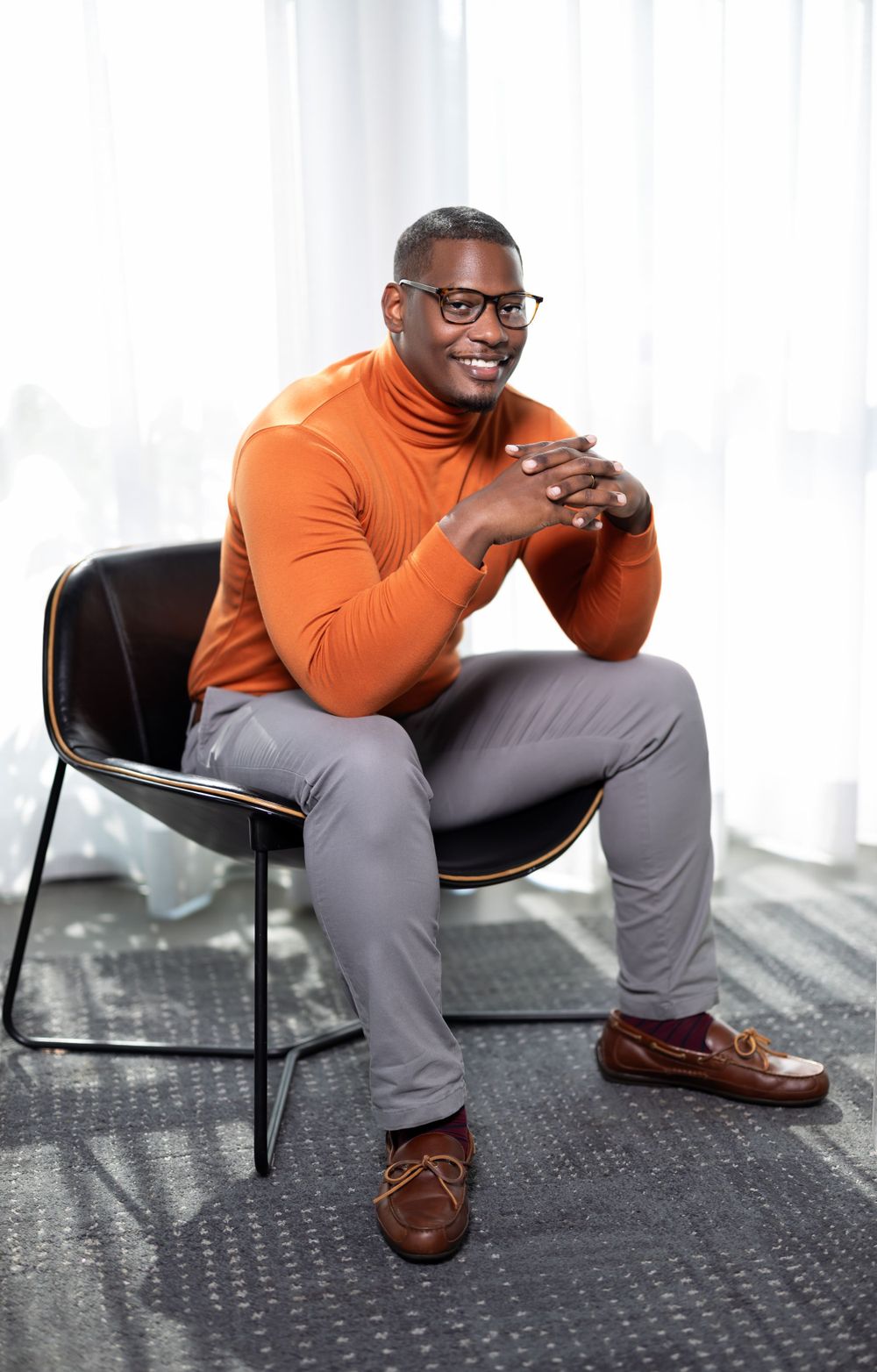 Professional wearing orange turtleneck and grey slacks seated in modern office chair.