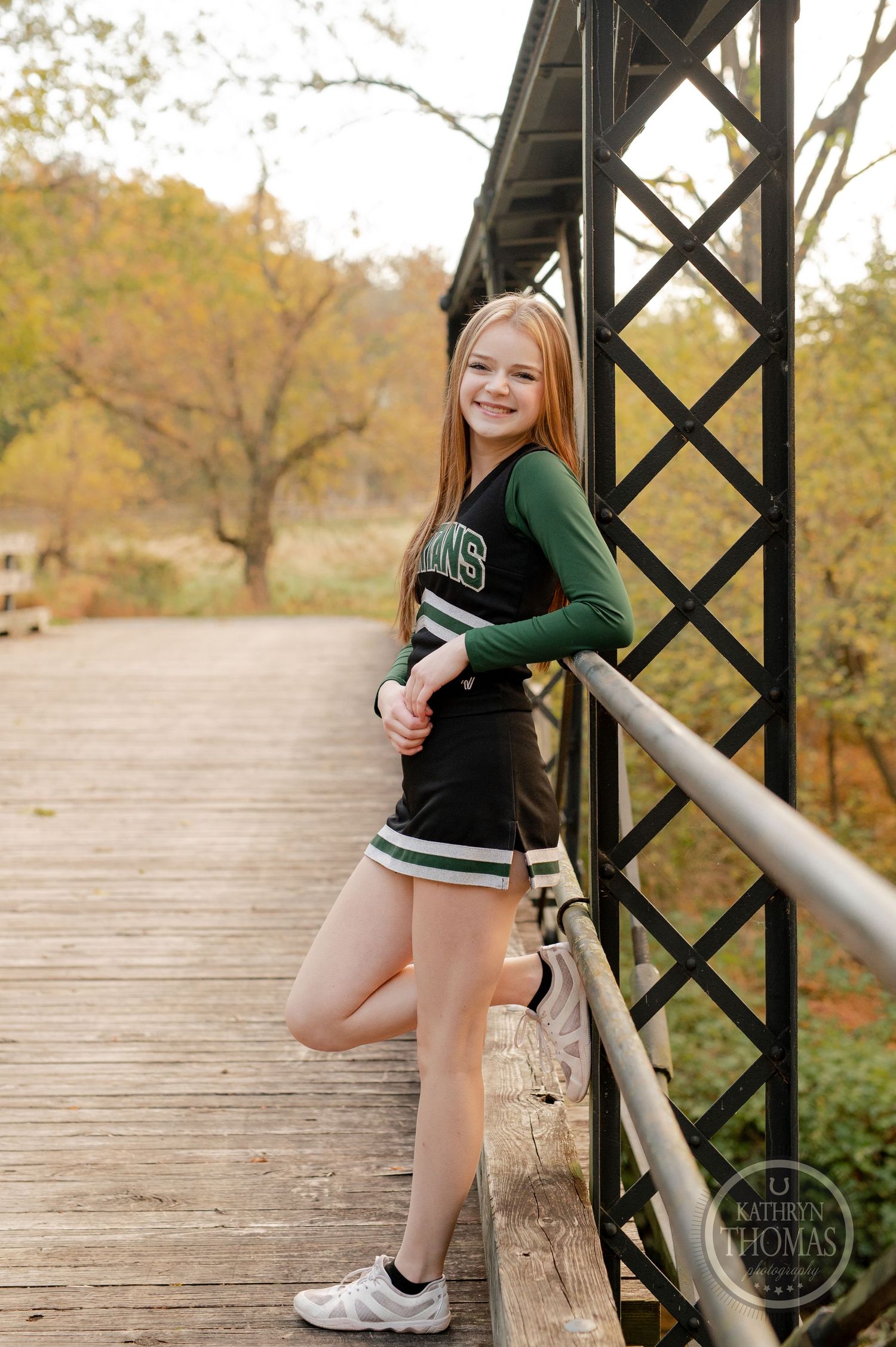Cheerleader in green uniform leans against metal bridge railing during fall photo session.