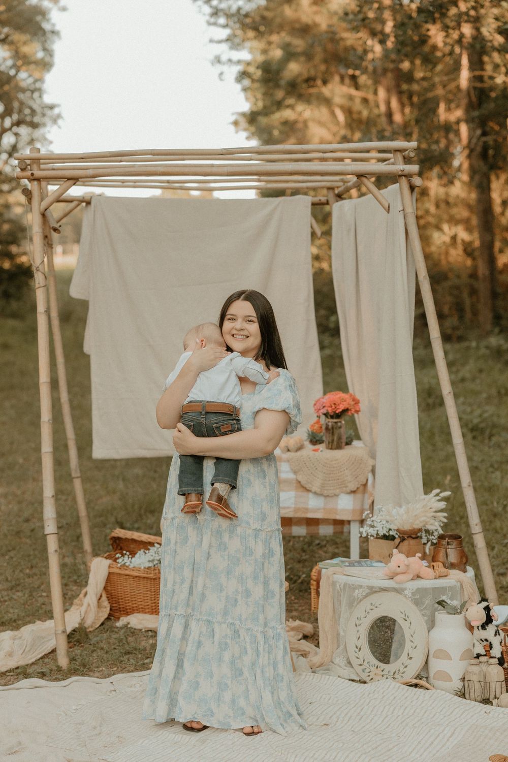 Woman in light blue floral maxi dress stands by rustic wooden display with baskets and vintage decor in outdoor setting.