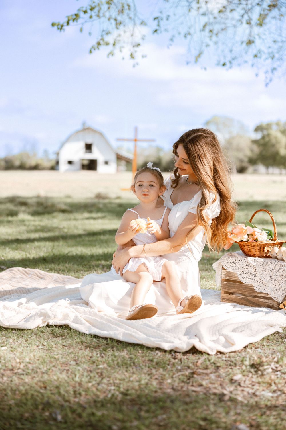 Mother and daughter in matching pink dresses enjoying a sunny picnic on a blanket near a white barn.