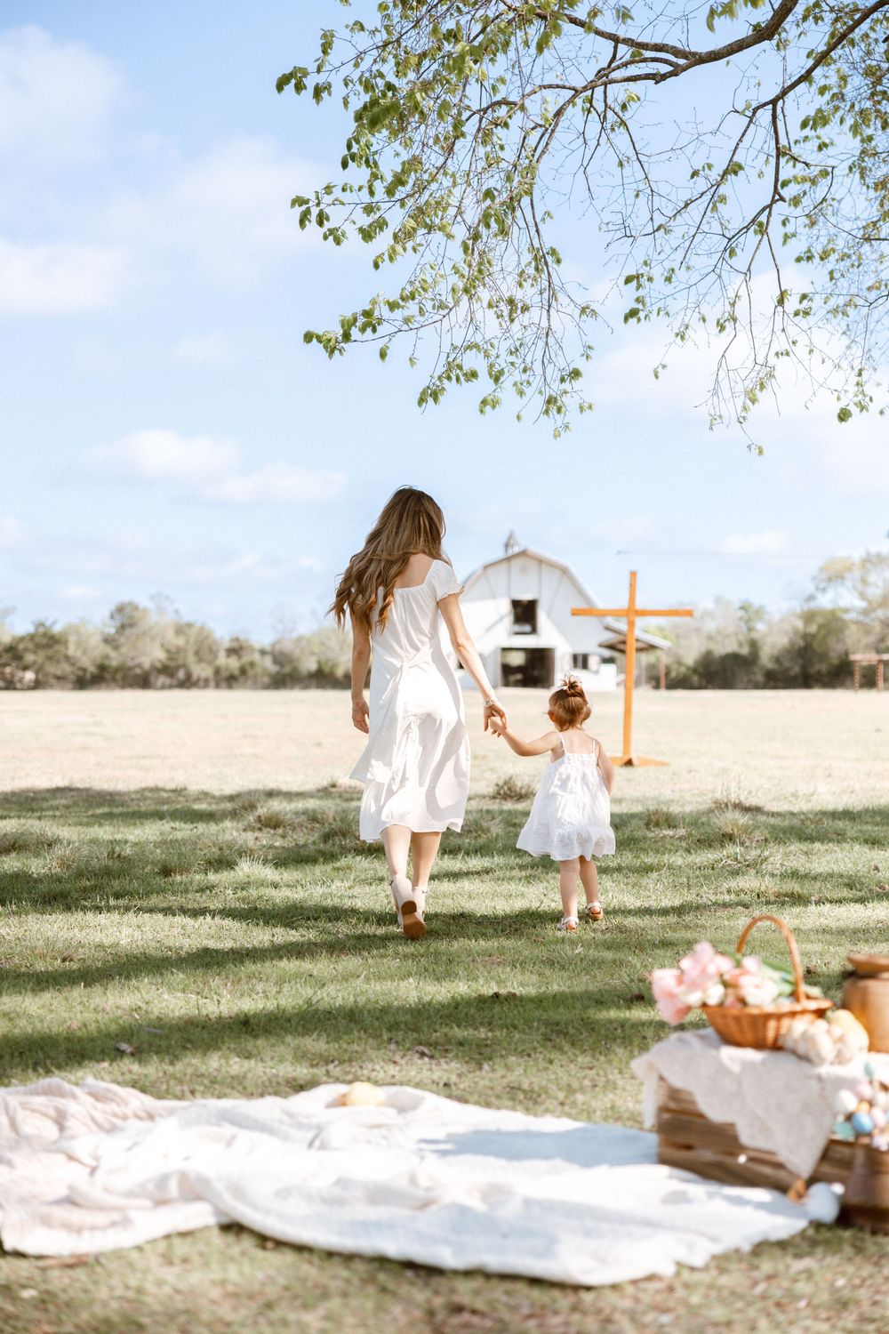 A mother and toddler in white dresses walk hand-in-hand across a sunny green field toward a white barn.