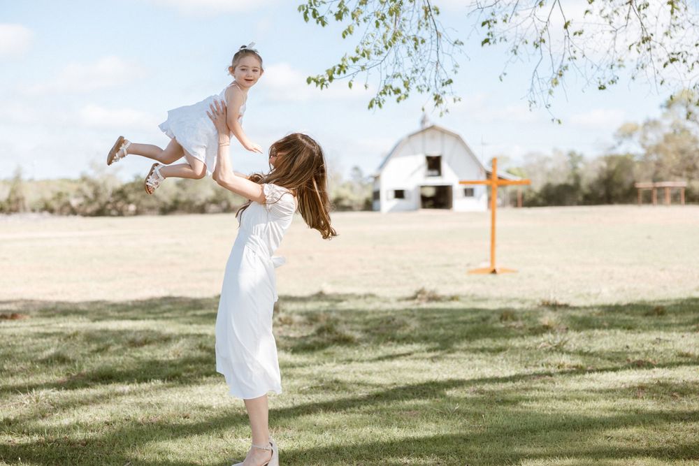Woman in white dress lifting a young girl in a sunny farm field with a white barn in the background.