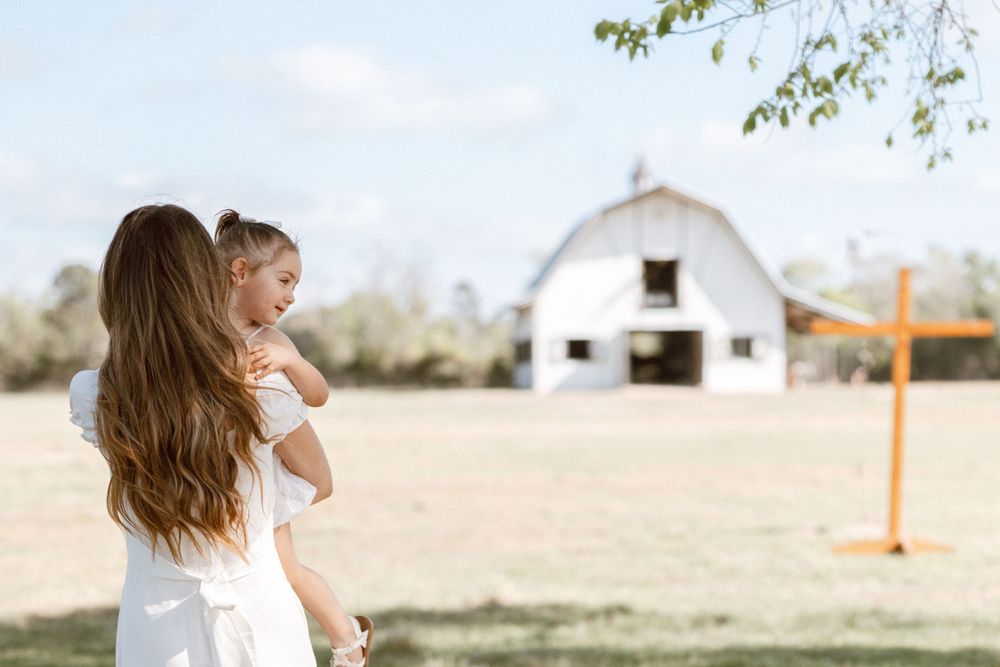 A mother holds her toddler while gazing at a white barn on a sunny farm, both dressed in white.