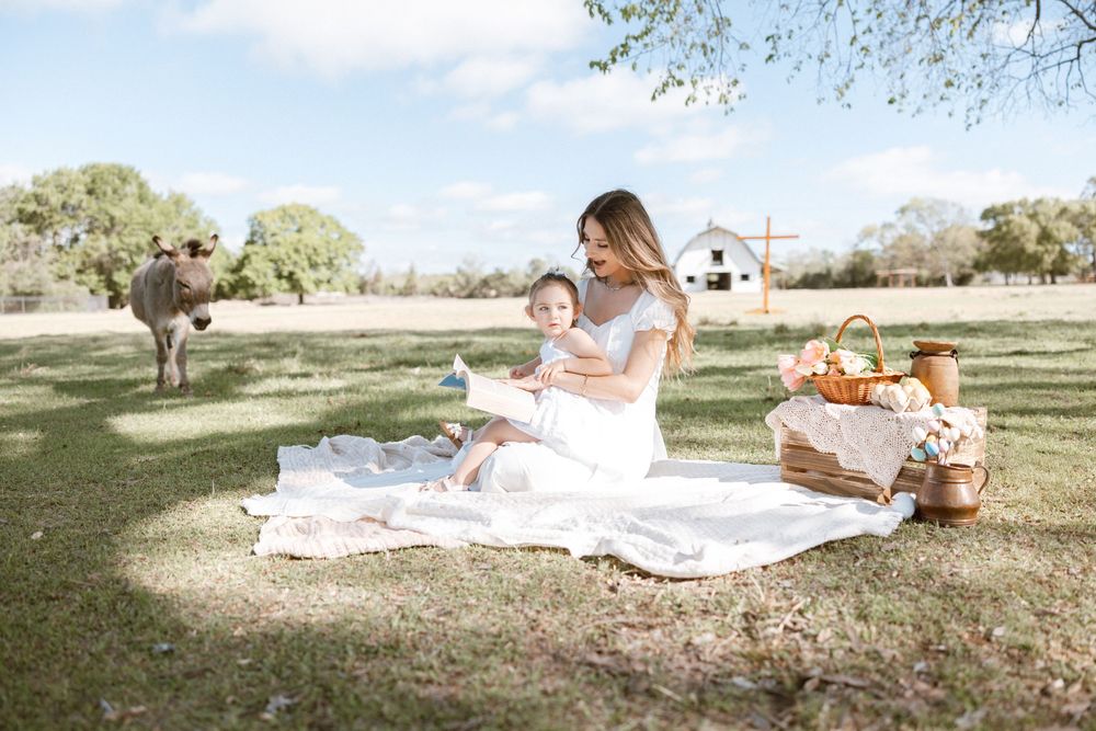 Mother and daughter in white dresses enjoying a picnic on green grass with a barn and cow in the background.