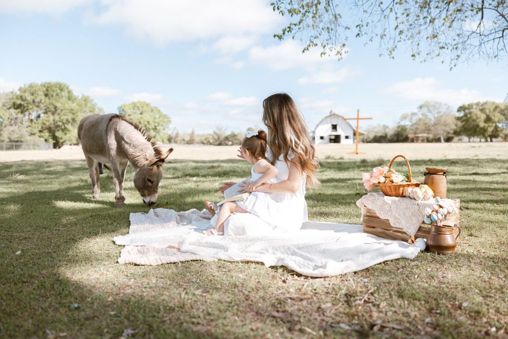 A mother and daughter in white dresses enjoy a picnic on a blanket near a grazing donkey on a sunny farm.
