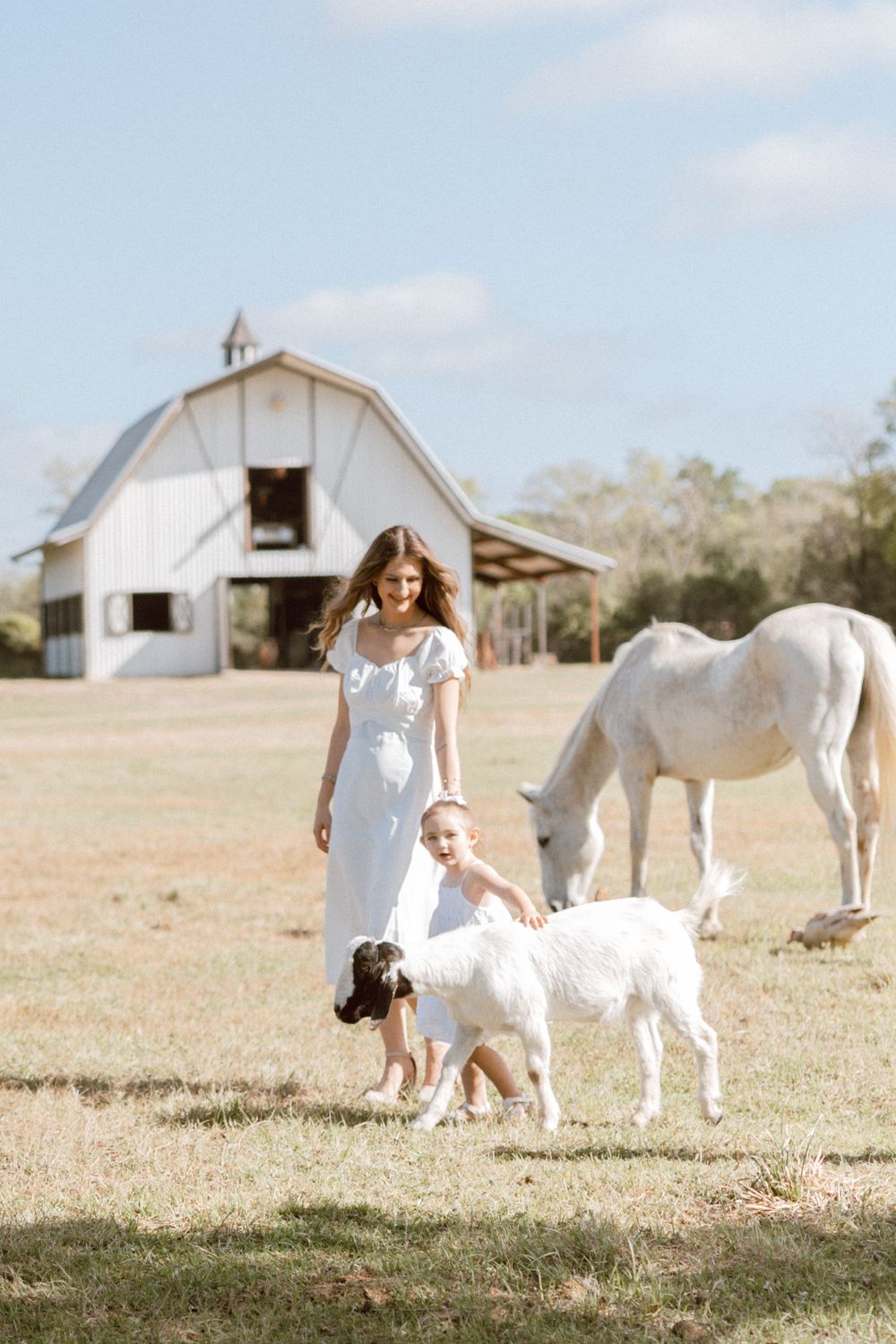 Woman in white dress with baby and goats on sunny farm, white horse and barn in background.
