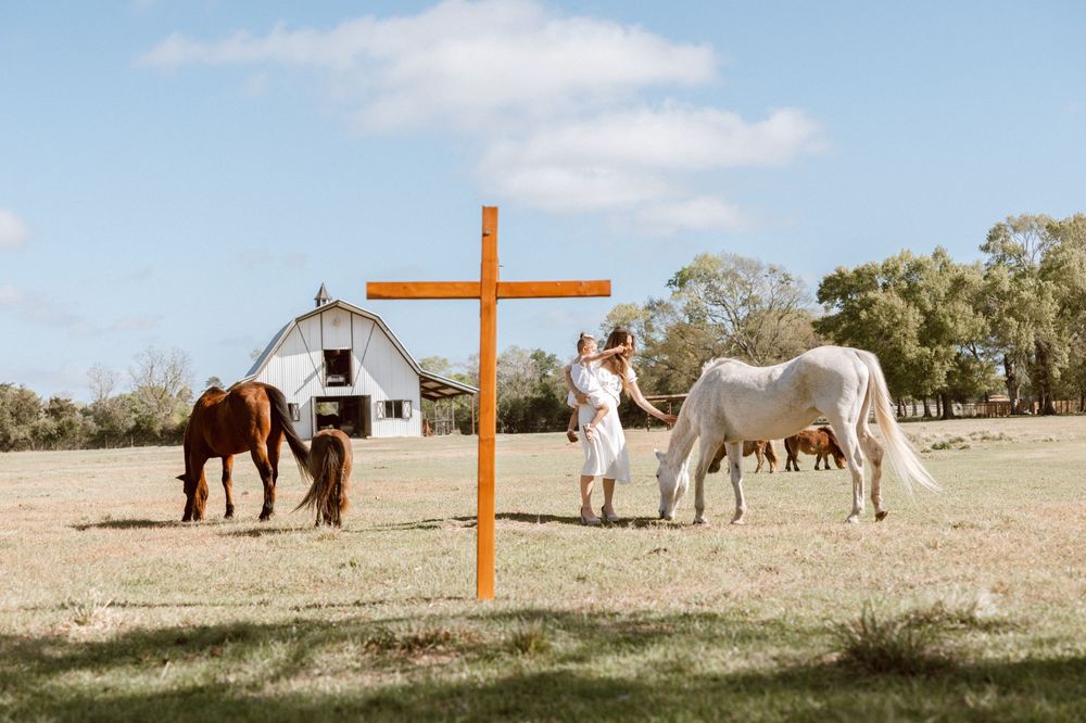 Couple stands near wooden cross on farm with white barn and horses grazing under blue sky.