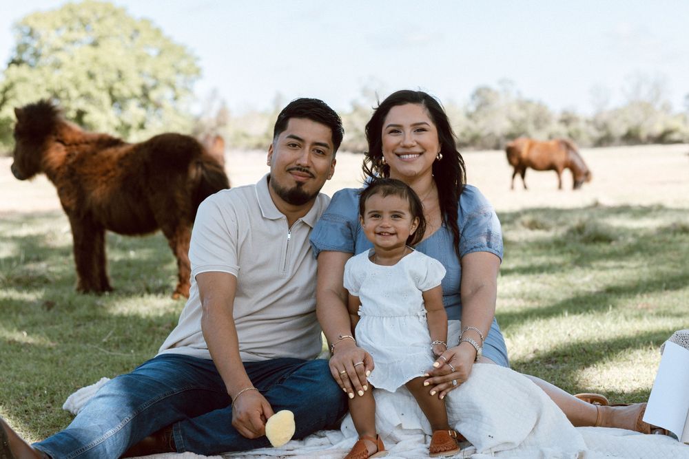 Family of three smiling on a picnic blanket outdoors with horses grazing in the background on a sunny day.