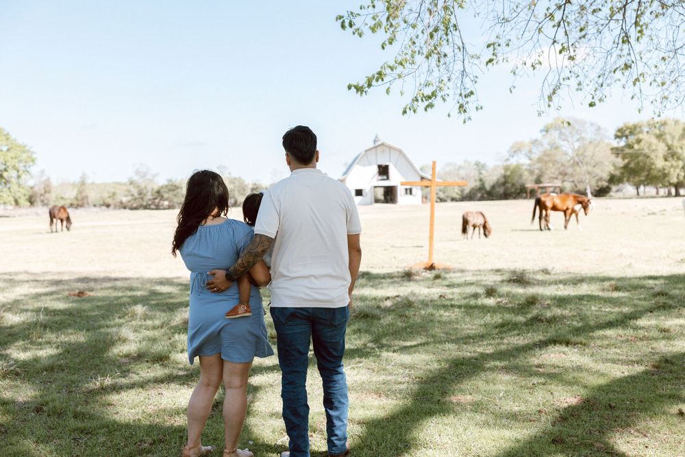 Family of three stands together viewing horses grazing near a white barn on a sunny farm day.