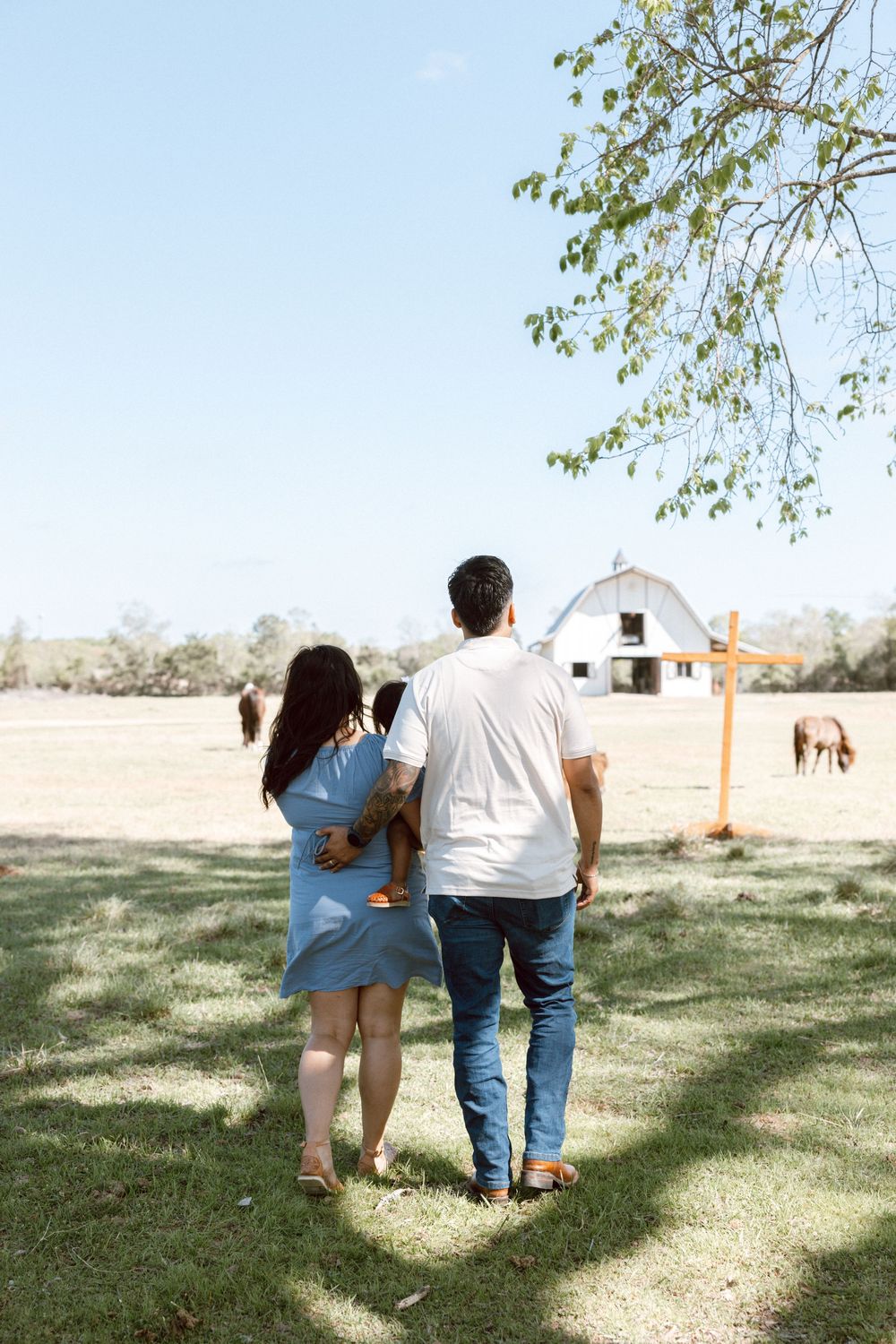 A family of three walks toward a white barn on a sunny farm, viewed from behind, with horses grazing in the background.