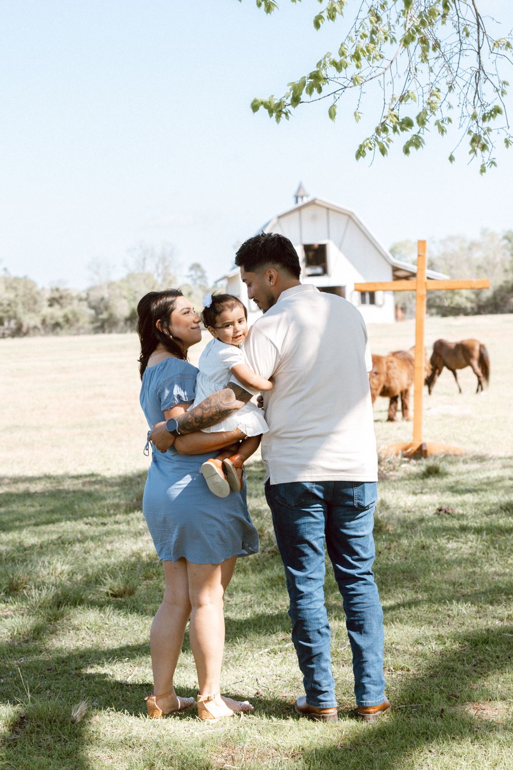 A young family with a baby poses on a sunny farm with a white barn and horses in the background.