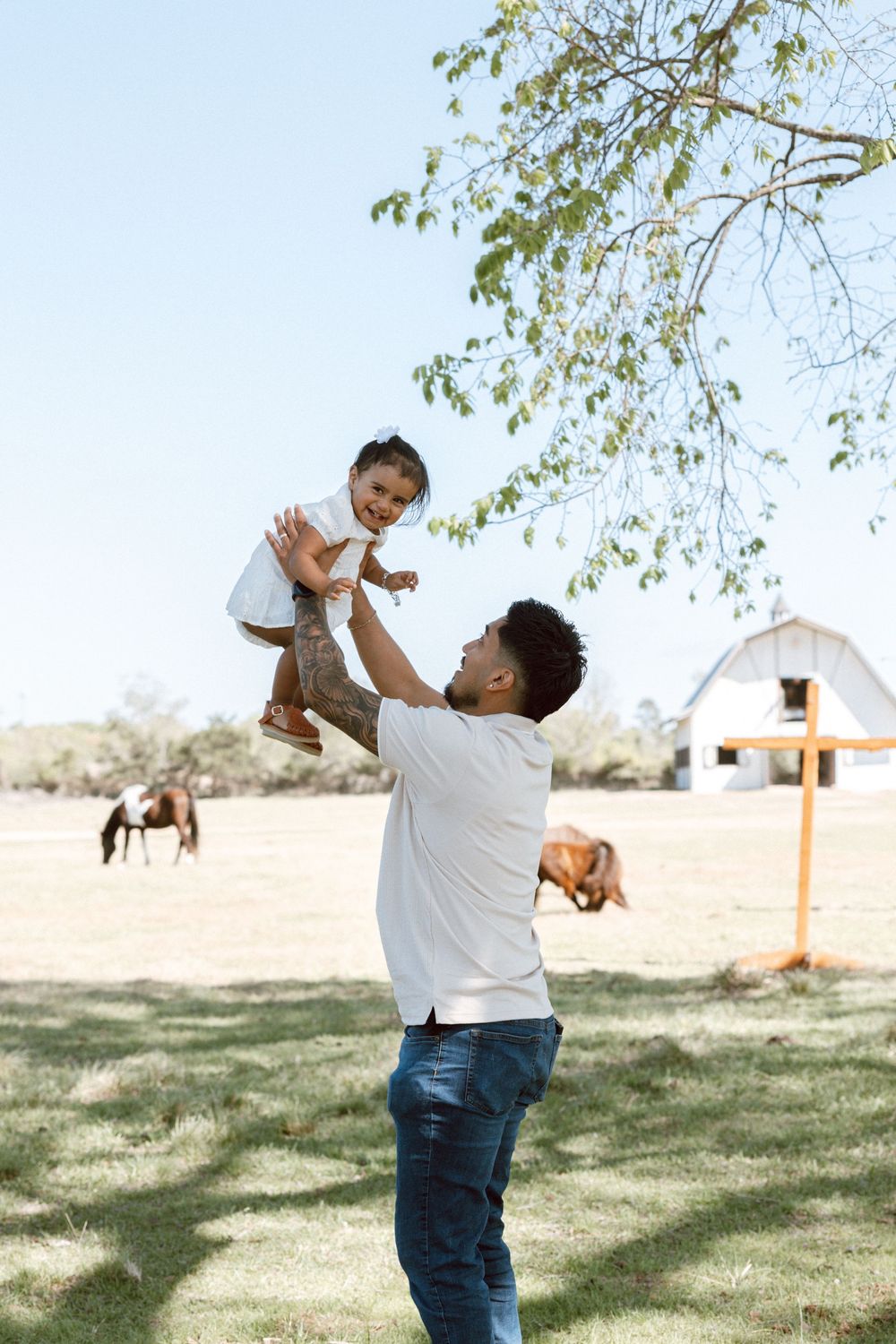 Father joyfully lifting smiling child in the air on a sunny farm with horses and white barn in background.