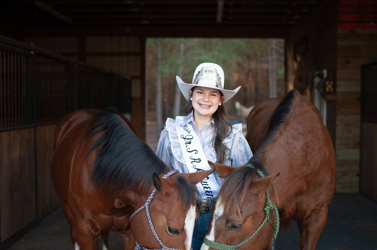 A pageant winner in white cowboy hat poses with two brown horses in a stable setting.