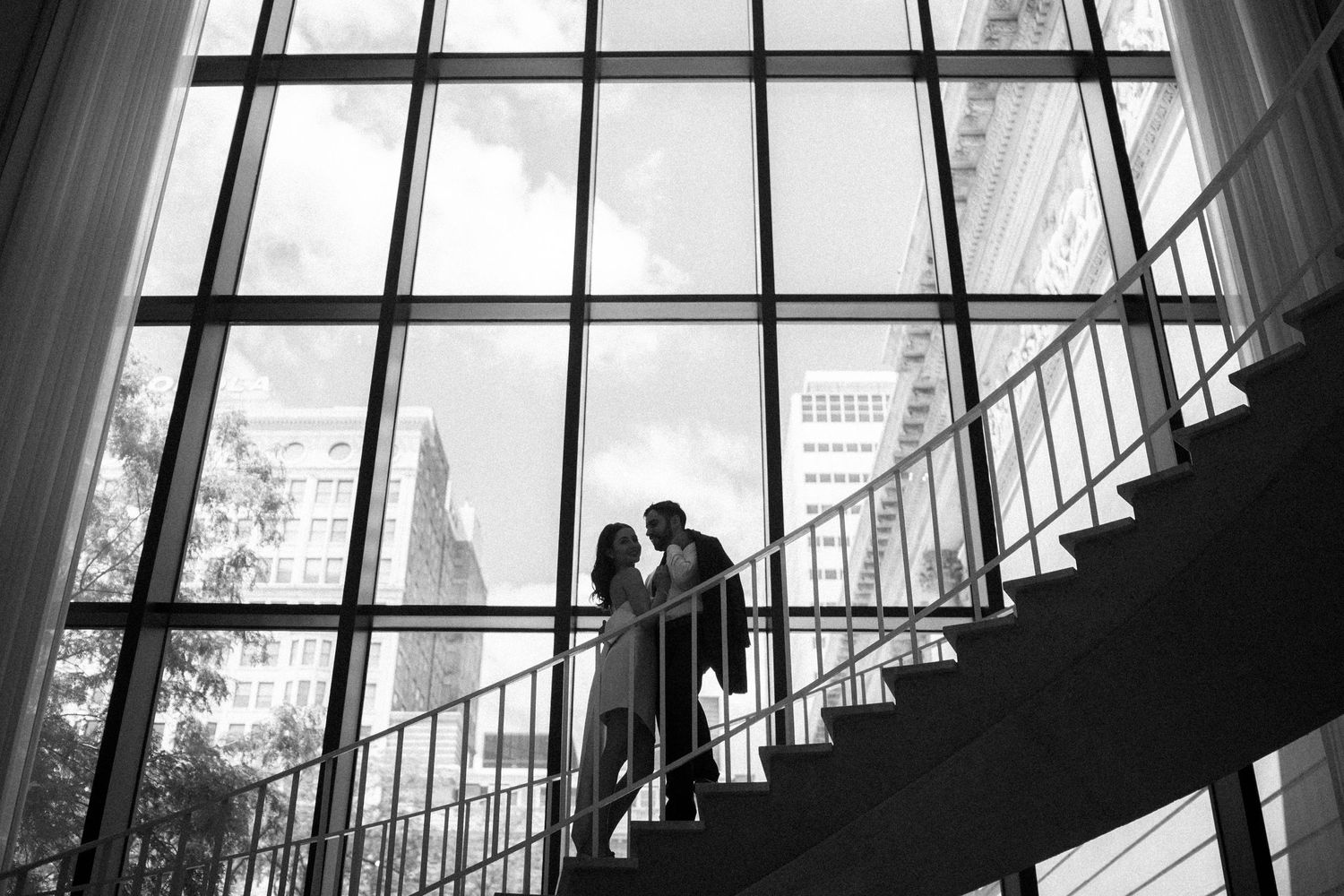 Dramatic silhouette of couple sharing a romantic moment on staircase against large industrial windows in black and white.