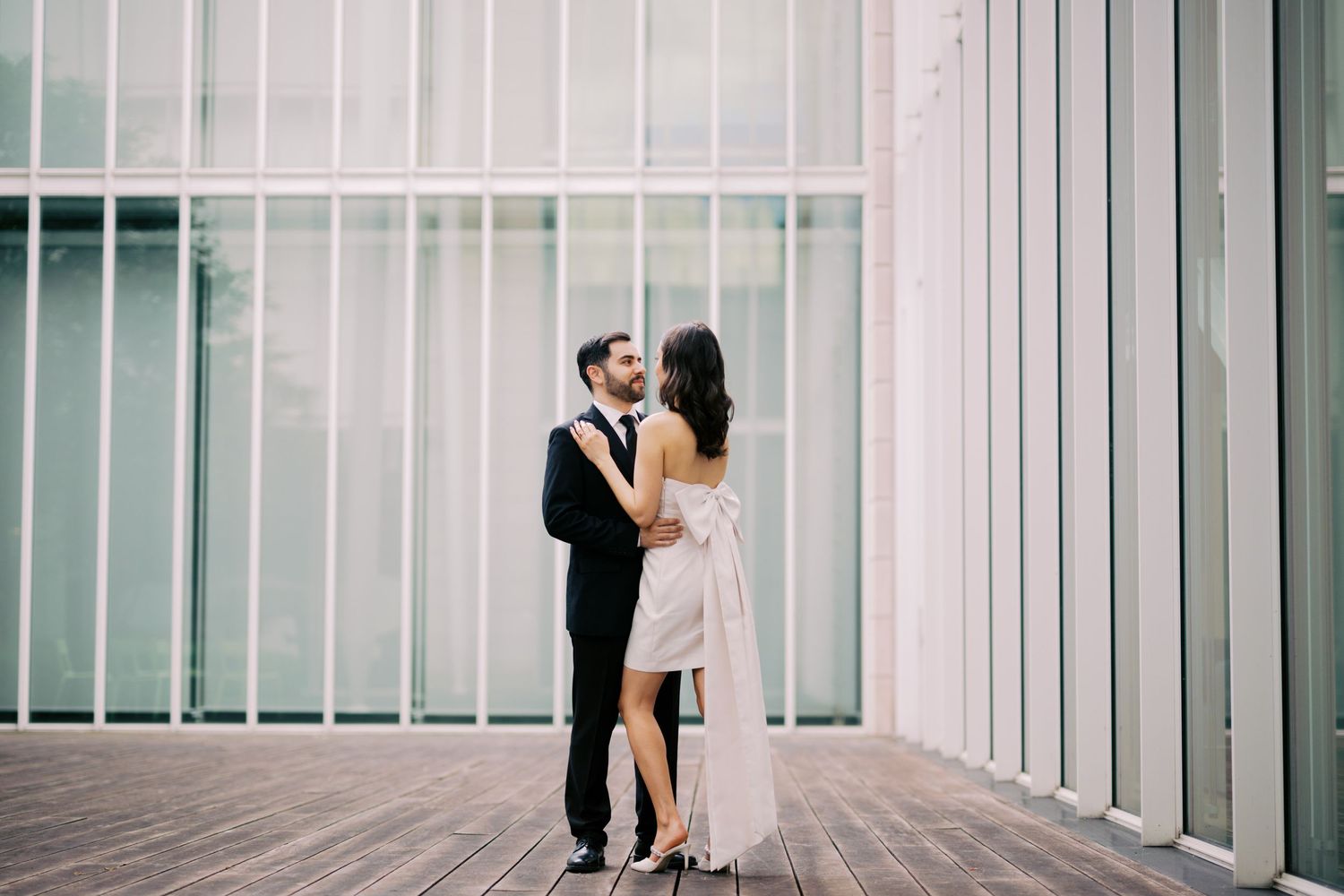 A couple in formal attire embrace on a modern balcony with white architectural details in the background.