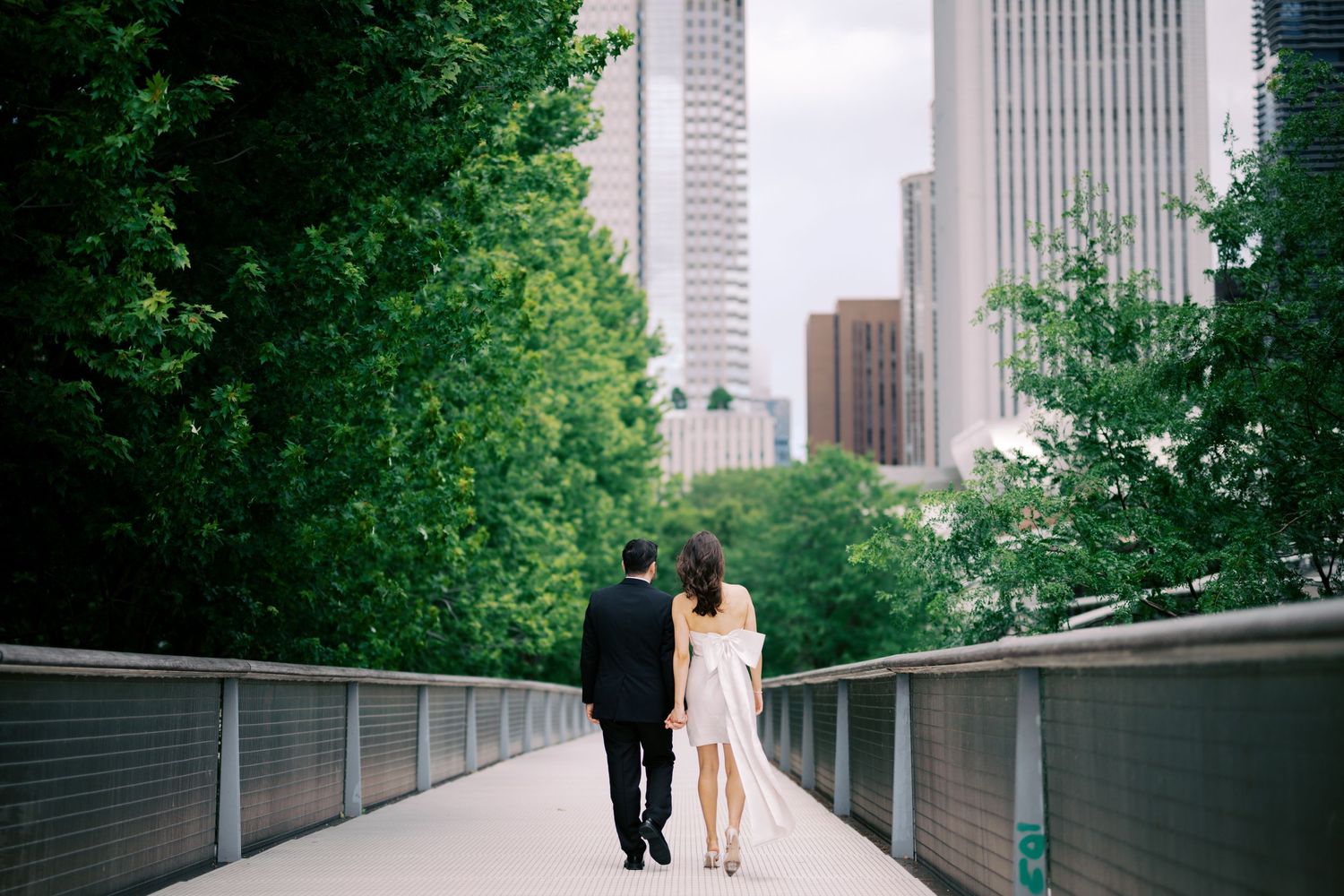 A couple walks together on an elevated path surrounded by green trees and urban skyscrapers in a city setting.