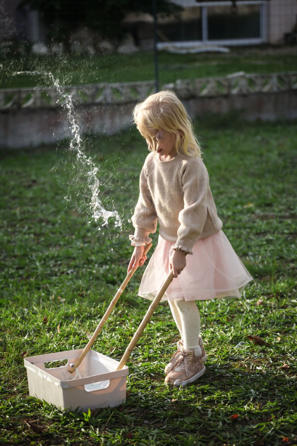 Young child in pink dress playing with toy cleaning set in backyard.