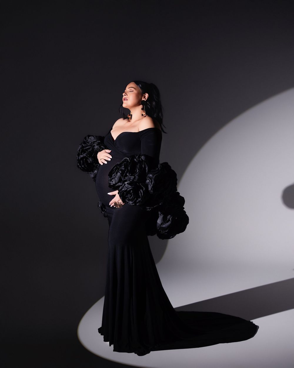 Woman in elegant black off-shoulder gown with floral ruffle sleeves, posing dramatically in studio lighting.