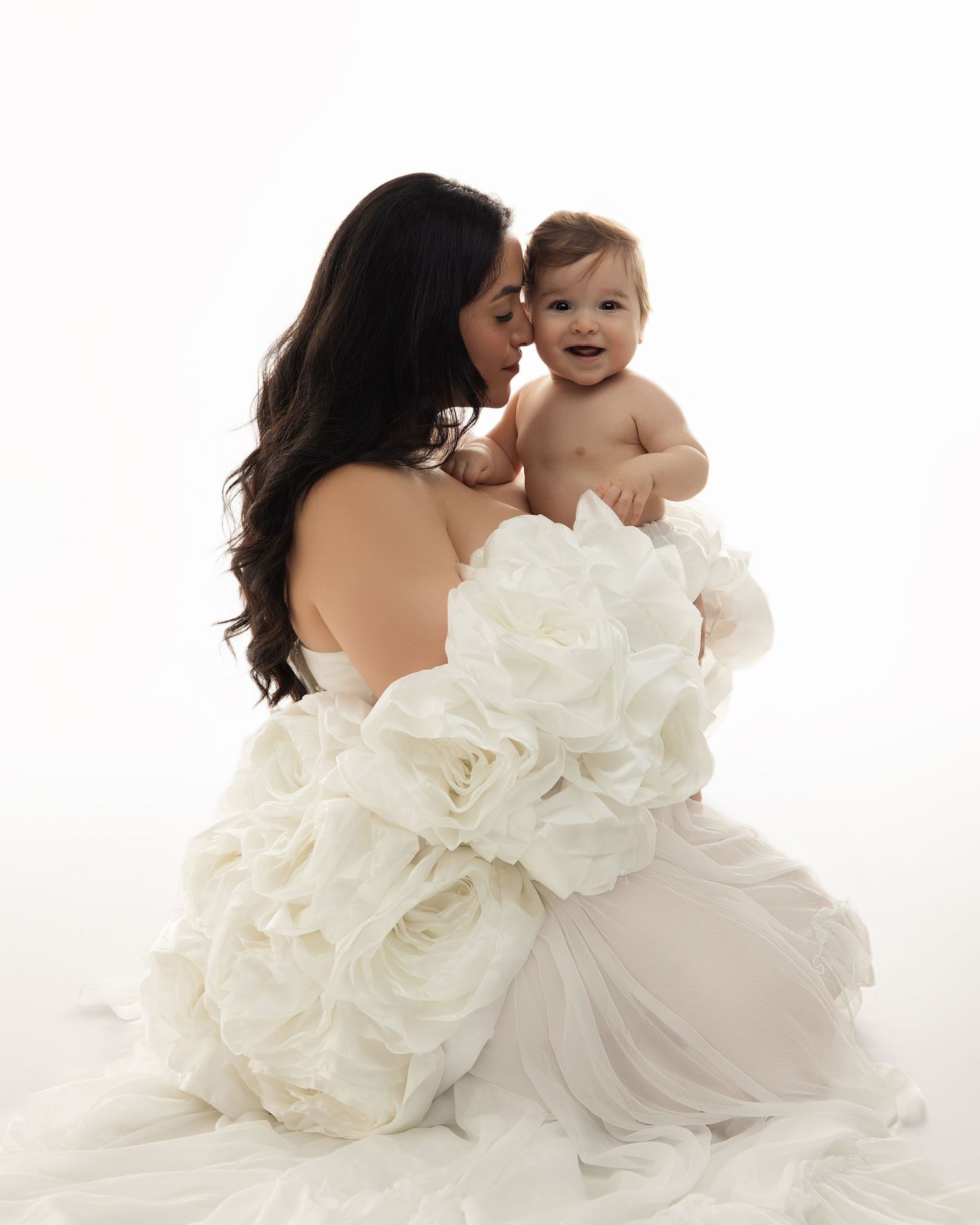 A parent in a flowing white gown holds a joyful baby against a bright white studio backdrop.