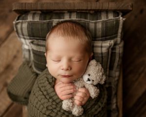 Sleeping newborn wrapped in knit sweater holding small teddy bear on plaid cushion.