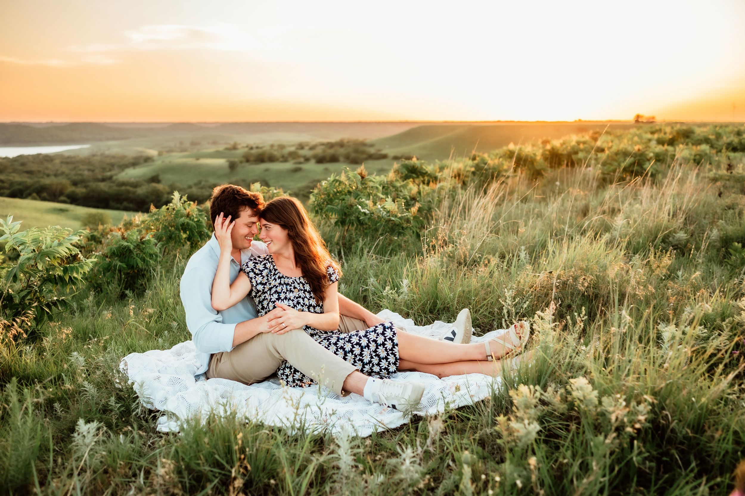 A couple relaxes together on a white blanket in a scenic grassy hilltop setting during golden hour.