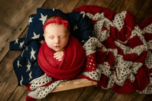 Newborn sleeping on American flag blanket wearing red knit wrap.
