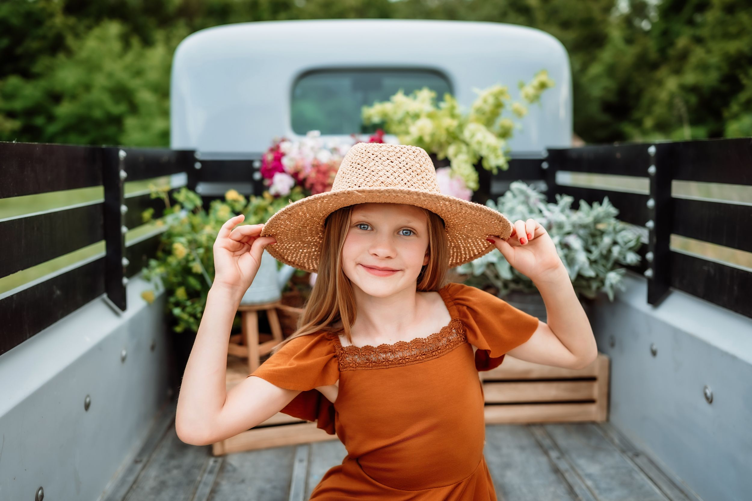 Rustic farm scene with straw hat and rust-colored dress in back of vintage truck with flower boxes and greenery.