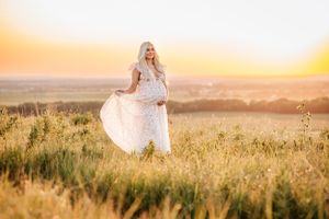White lace dress flows in golden sunlit meadow during sunset maternity photo session.