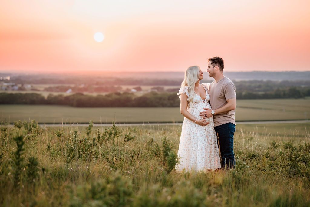 A couple embraces in a meadow during golden hour sunset against a rolling countryside landscape.