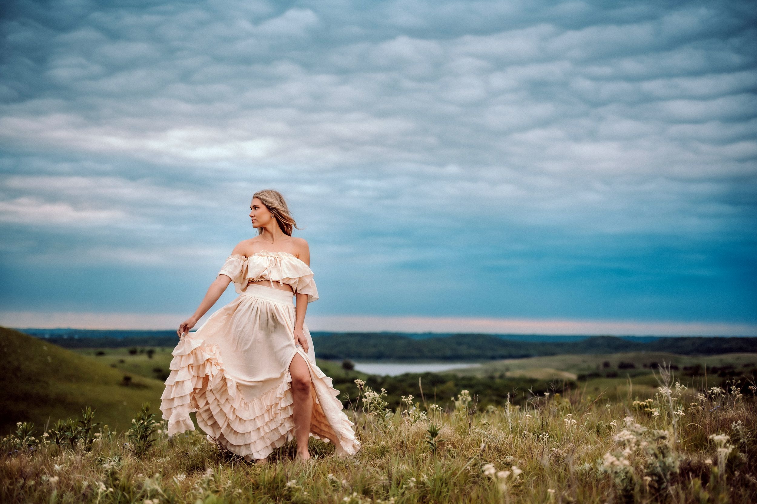 A person in a flowing blush pink dress stands gracefully on a hilltop overlooking a vast coastal landscape at sunset.