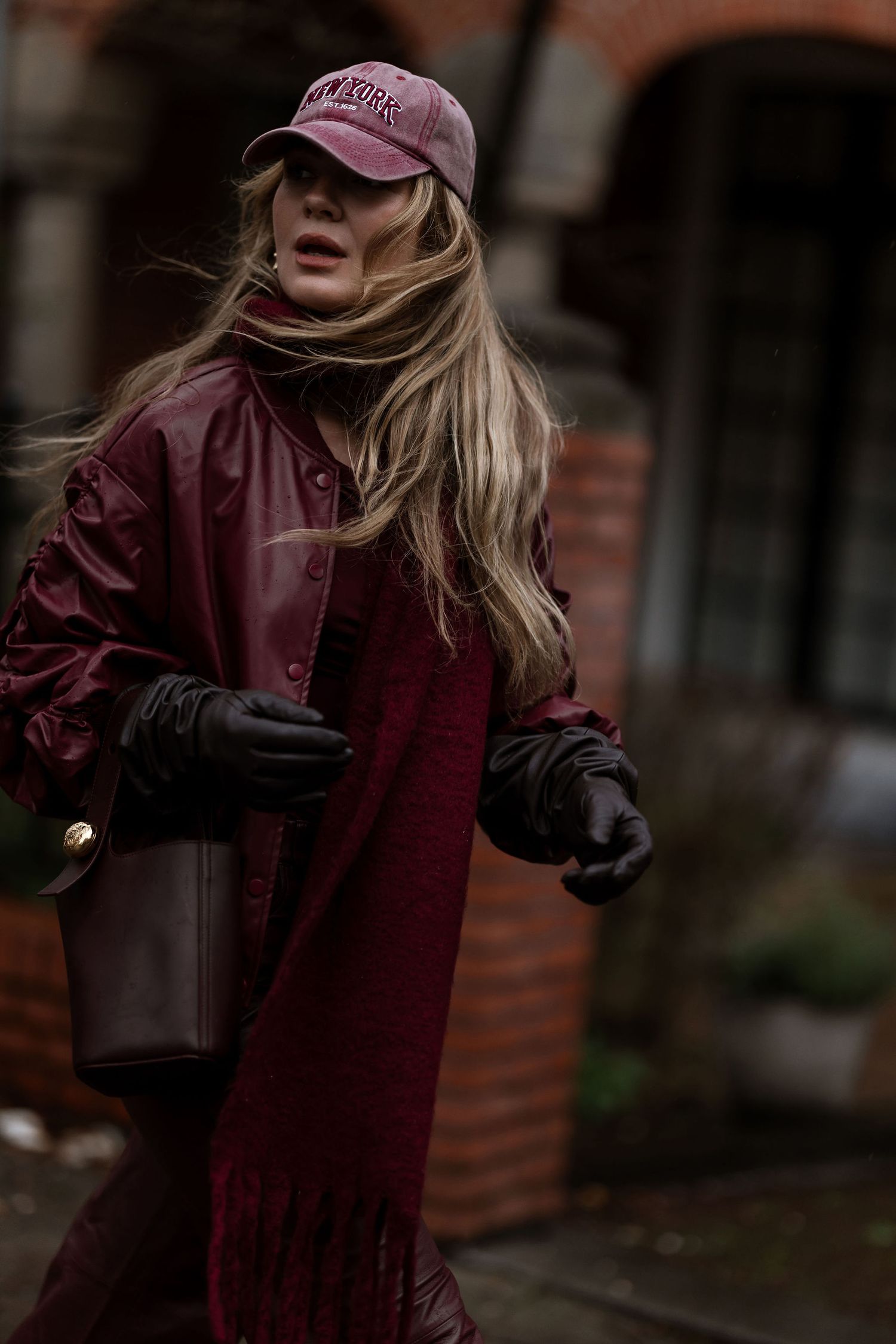 Street style look with burgundy coat, baseball cap, and leather bag on a moody autumn day.