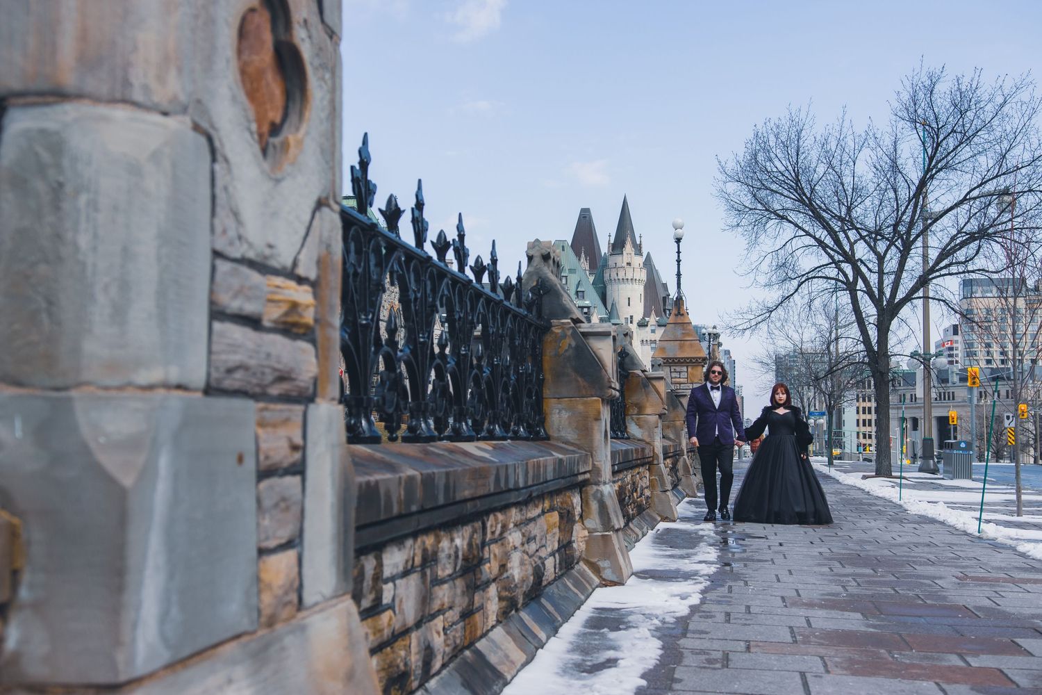 Winter scene of pedestrians walking along a snowy sidewalk beside ornate iron gates and stone walls.