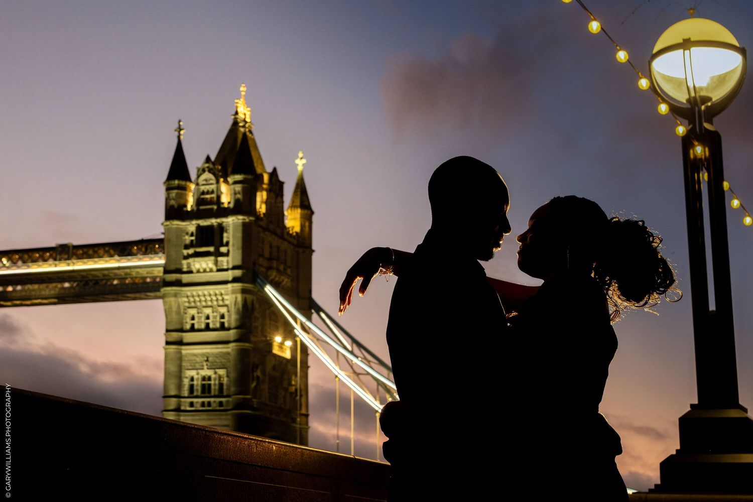 A silhouetted couple stands near a lamp post against the illuminated Tower Bridge in London at sunset.